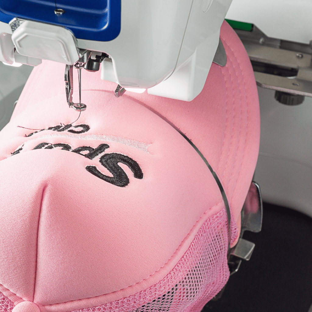 Close-up of embroidery machine stitching a black logo on a pink cap, demonstrating precision garment branding.